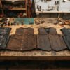 Wide shot of a leather workshop table with several finished leather MC vests laid out side by side — black cowhide, brown buffalo, and a lambskin piece — showing texture variety. Overhead or 45-degree angle, warm workshop lighting, visible stitching detail. Should feel authentic and hands-on, not stock-photo staged.