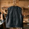Back view of a plain, unpatched leather MC vest hanging on a wooden hanger, showing the full uninterrupted back panel with visible grain and no center seam. Studio or workshop lighting, back-facing angle.