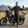 A rider standing beside a motorcycle wearing a well-fitted black full-grain leather MC vest, front zipped, arms slightly raised showing full mobility. Warm, natural daylight, garage or open-road backdrop. Landscape orientation.